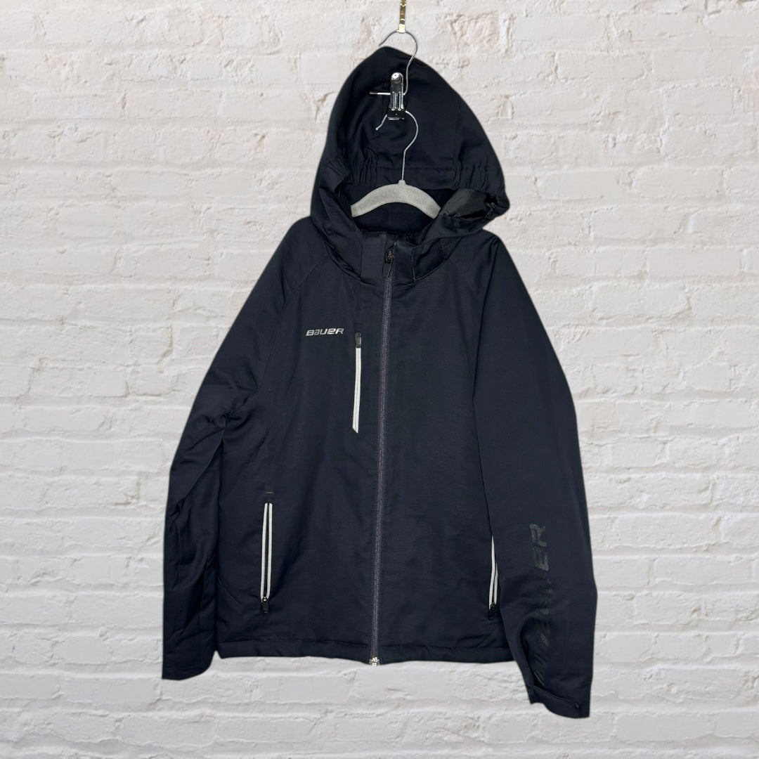 Black Bauer hooded windbreaker jacket with zipper pockets, hanging on a hanger against a brick wall.