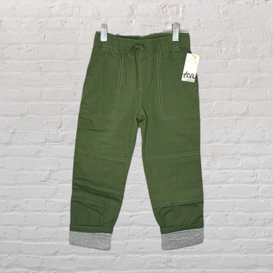 Children's green jogger pants with elastic waist and grey cuffs, hanging with a tag from Tea brand.