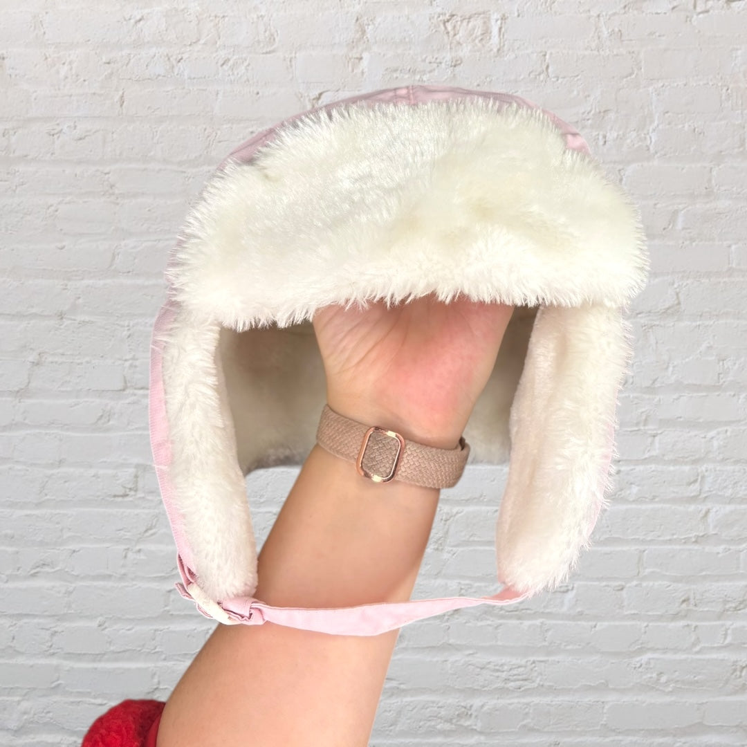 Pink trapper hat with white faux fur lining and ear flaps against a white brick background.