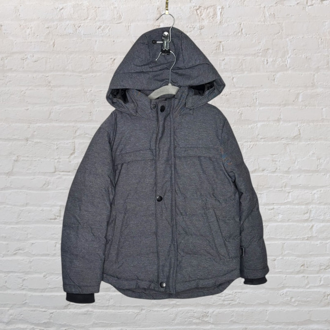 Charcoal grey hooded puffer jacket with button and zipper front, displayed against a white brick wall.
