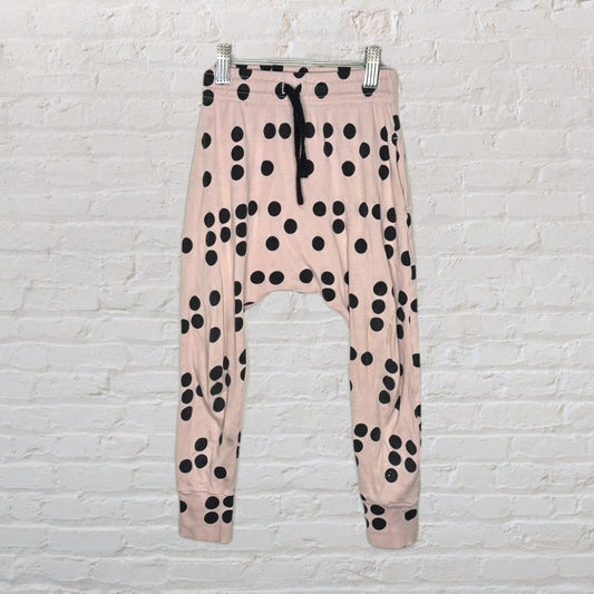 Pink drop-crotch pants with black polka dots, featuring an elastic waistband and drawstring, hanging against a brick wall.
