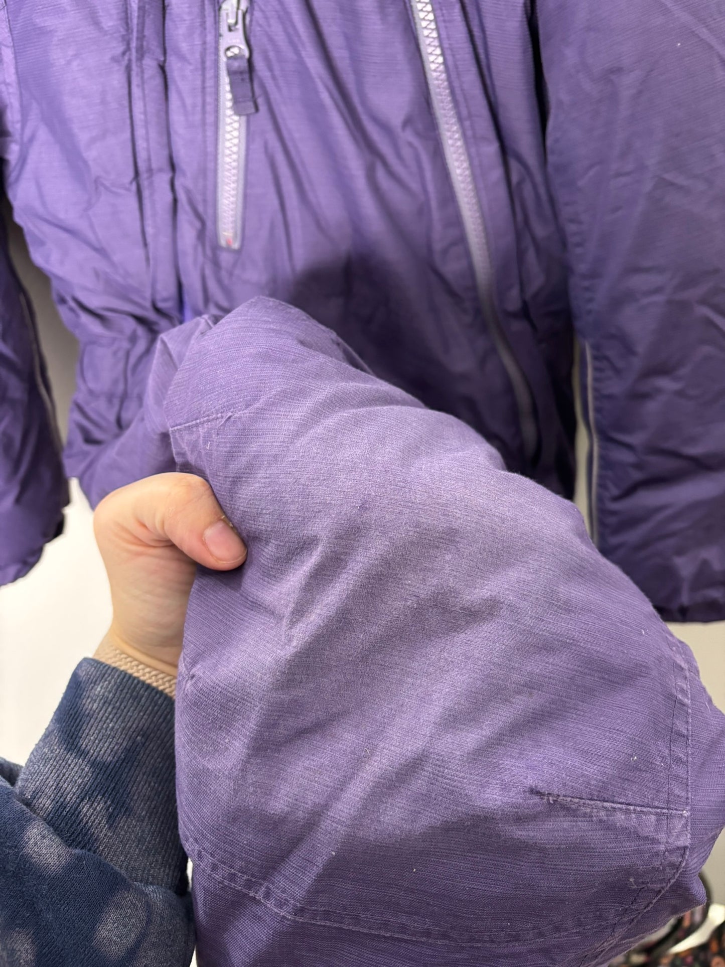 Purple insulated kids' snowsuit with hood and zip closure, hanging against a white brick wall.