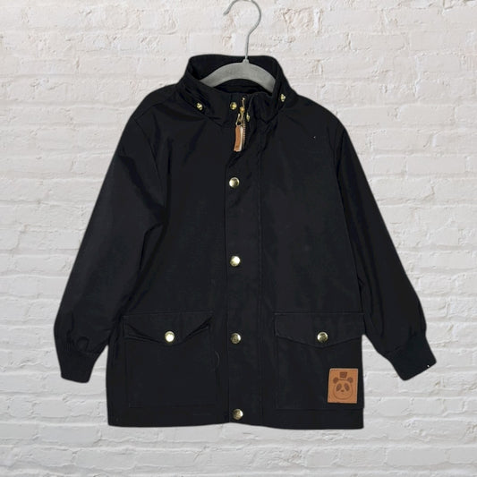 Kids black hooded parka jacket with gold buttons and front pockets, hanging on a hanger against a brick wall.