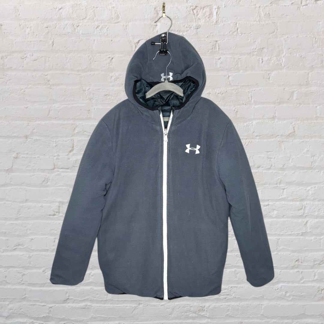 Camouflage patterned Under Armour puffer jacket with hood and white zipper, hanging on a hanger against a brick wall.