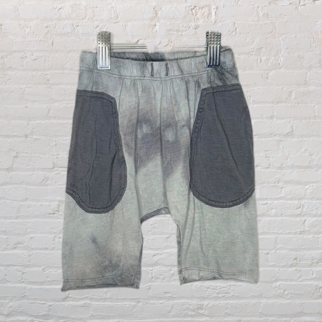 Grey shorts with large charcoal patch pockets, hanging on a hanger against a brick wall.