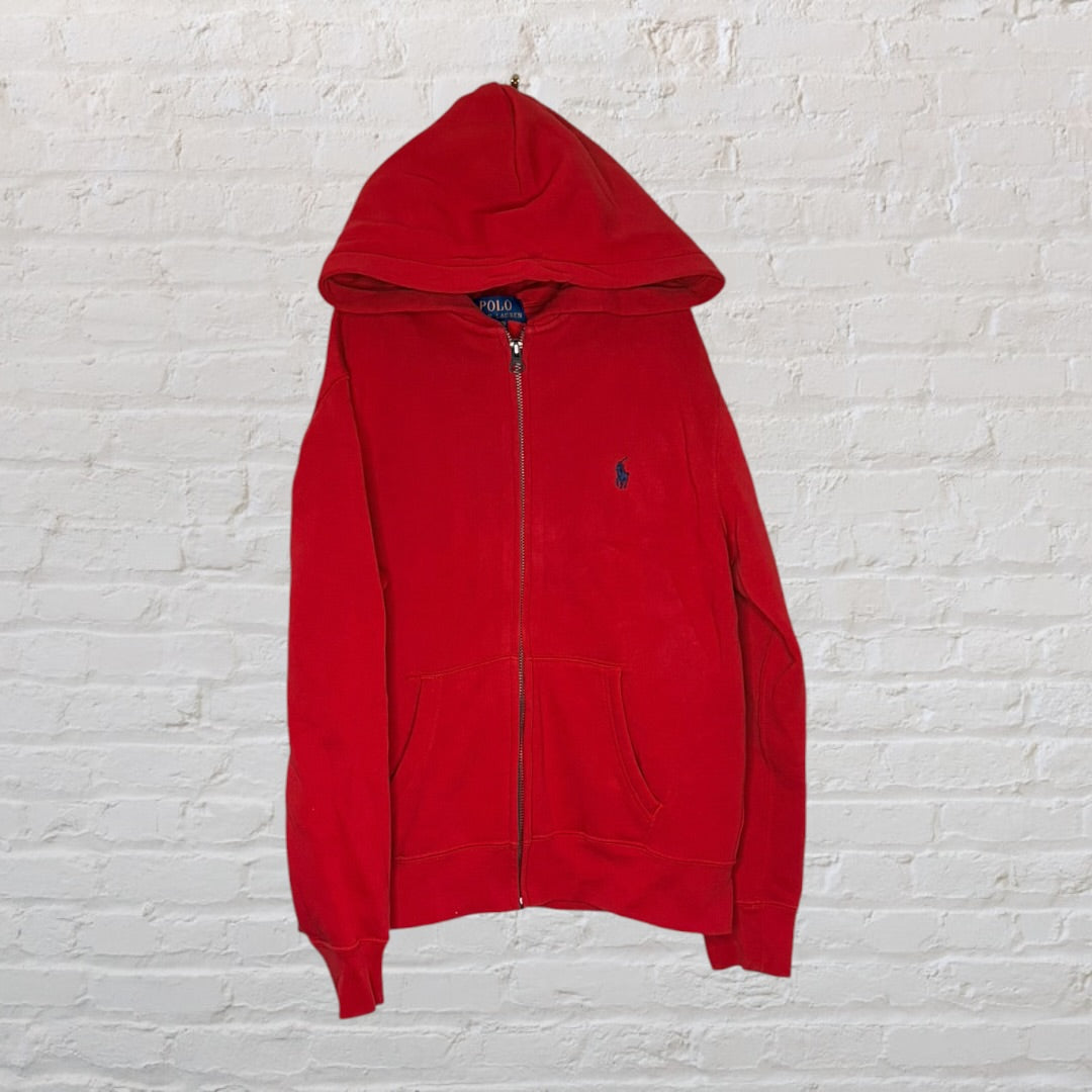 A red Polo Ralph Lauren zippered hoodie with a front pocket and embroidered logo, hanging against a white brick wall.