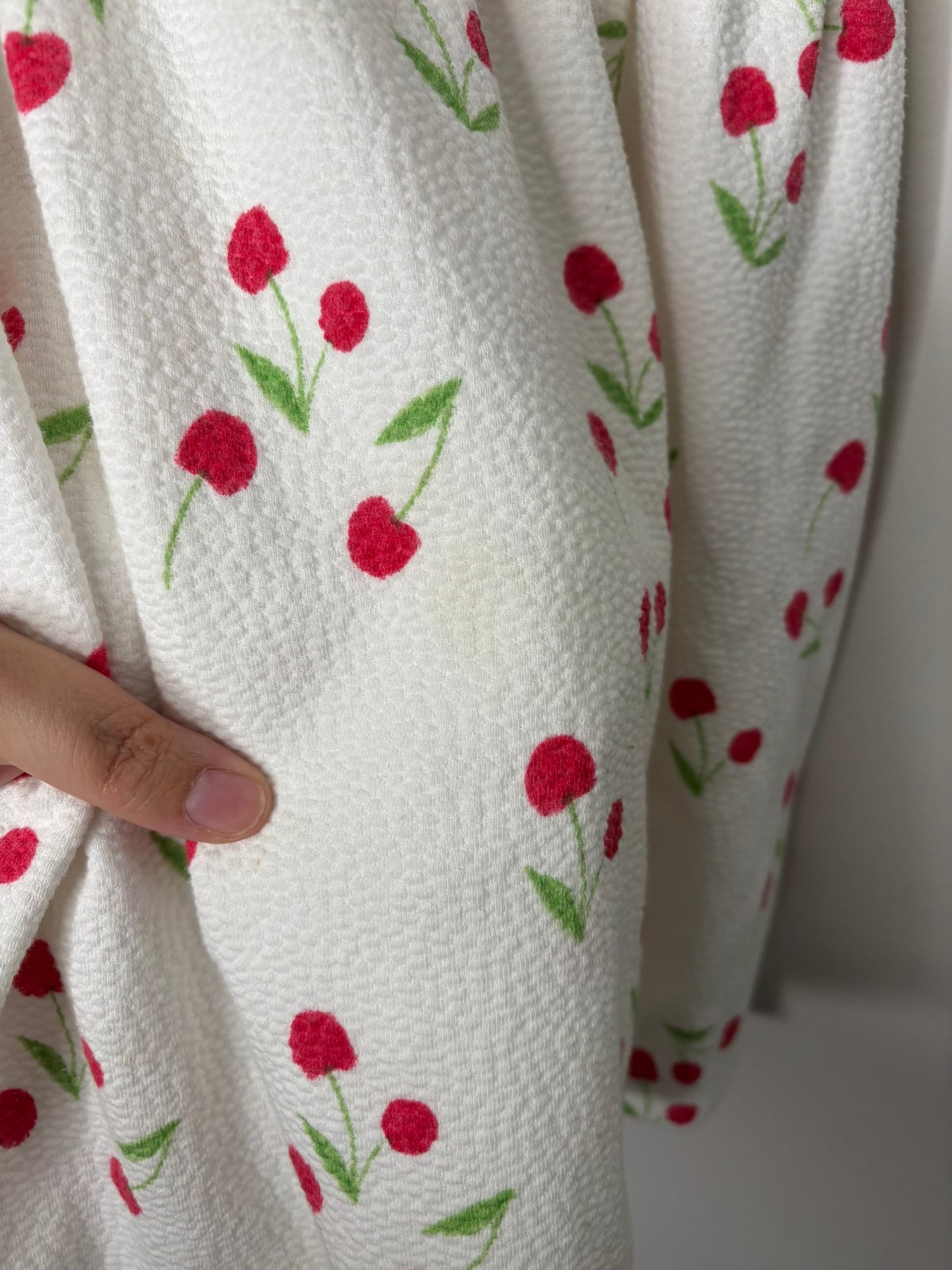 White dress with red cherry print and ruffled sleeves hanging on a black hanger against a textured wall.