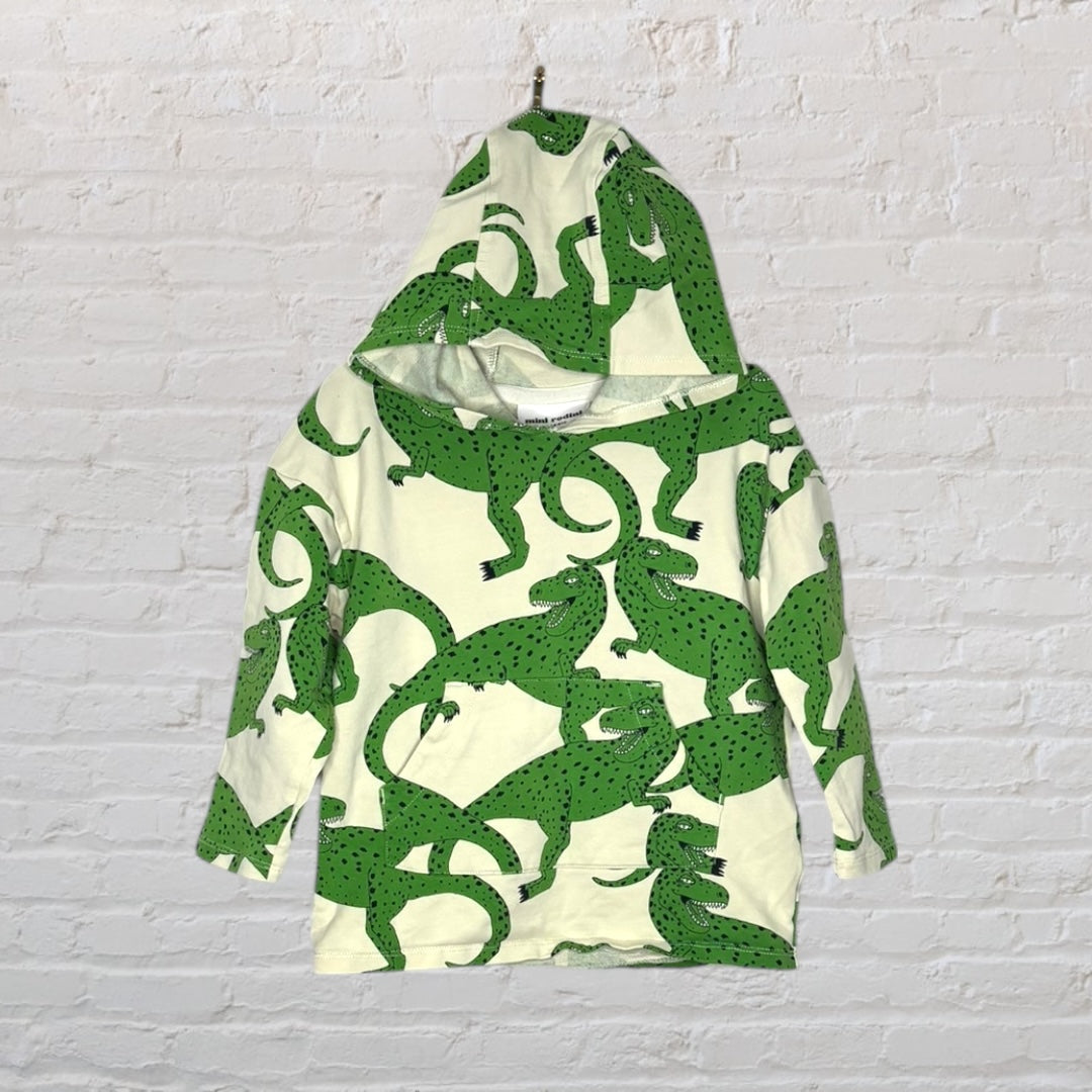 Kids' cream hoodie with green alligator print, featuring long sleeves and a hood, hanging against a white brick wall.