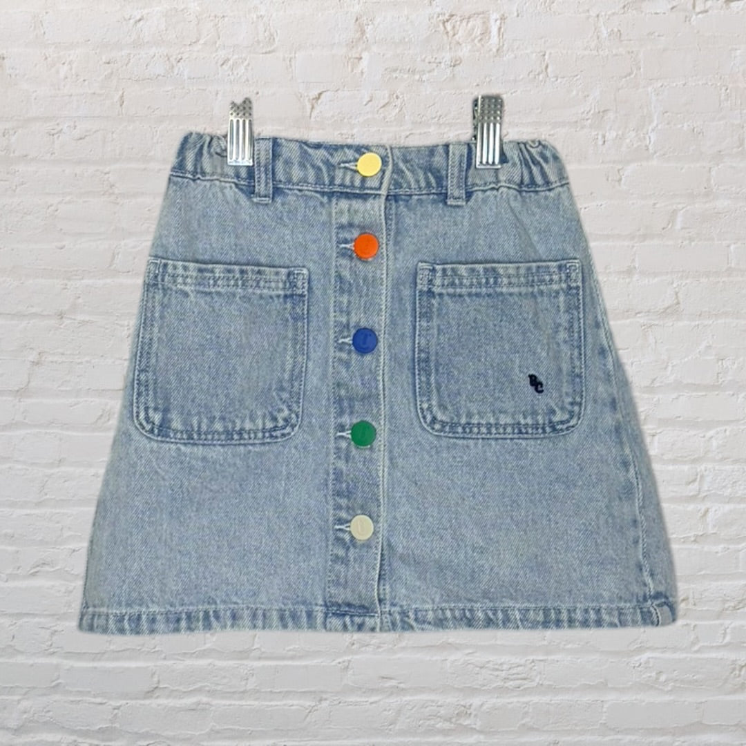 Light blue denim skirt with multicolored buttons and front pockets, displayed against a white brick background.