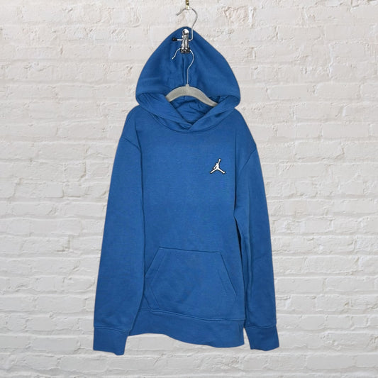 Blue Jordan hoodie with front pocket and embroidered logo, hanging against a white brick wall.