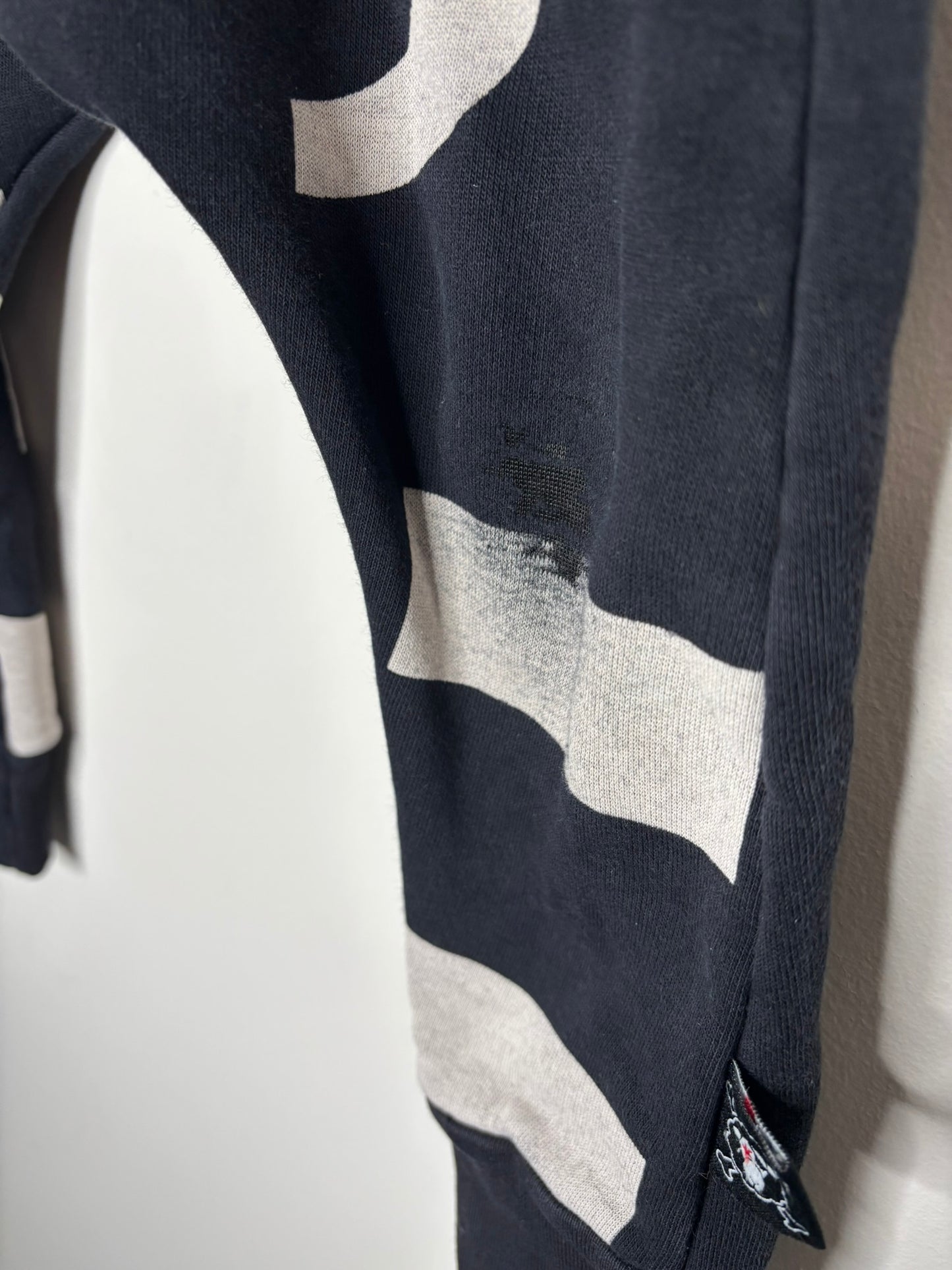 Black harem pants with bold white geometric patterns hanging on a hanger against a white brick wall.