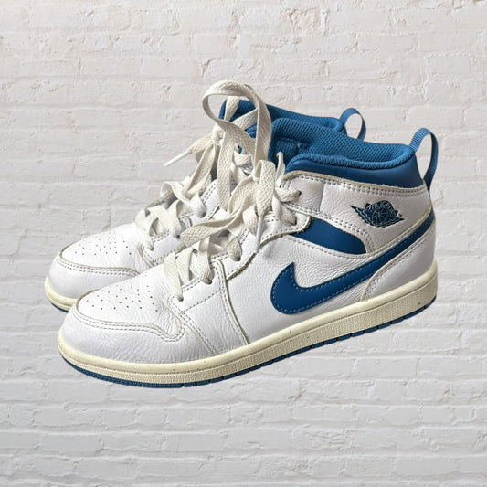 Pair of white and blue Nike Air Jordan Mid Sneakers with laces against a brick wall background.