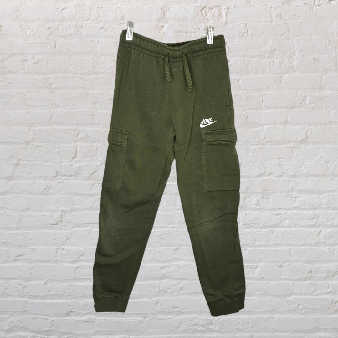 Olive green Nike cargo jogger pants with side pockets and drawstring waist, displayed hanging against a brick wall.