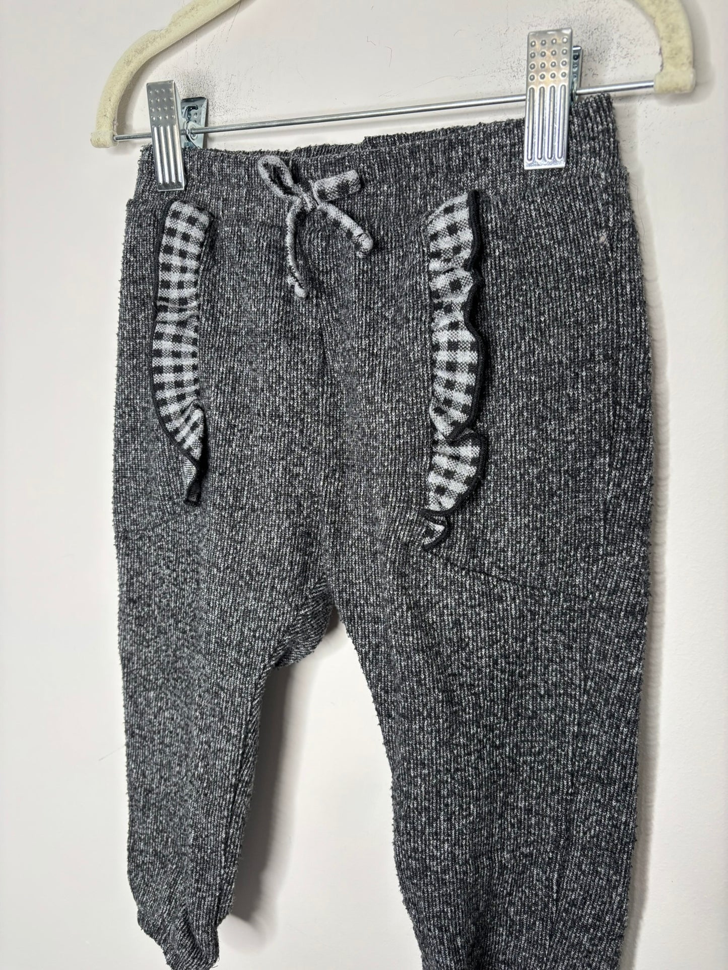 Charcoal knit pants with black and white gingham ruffle details, hanging against a white brick wall.