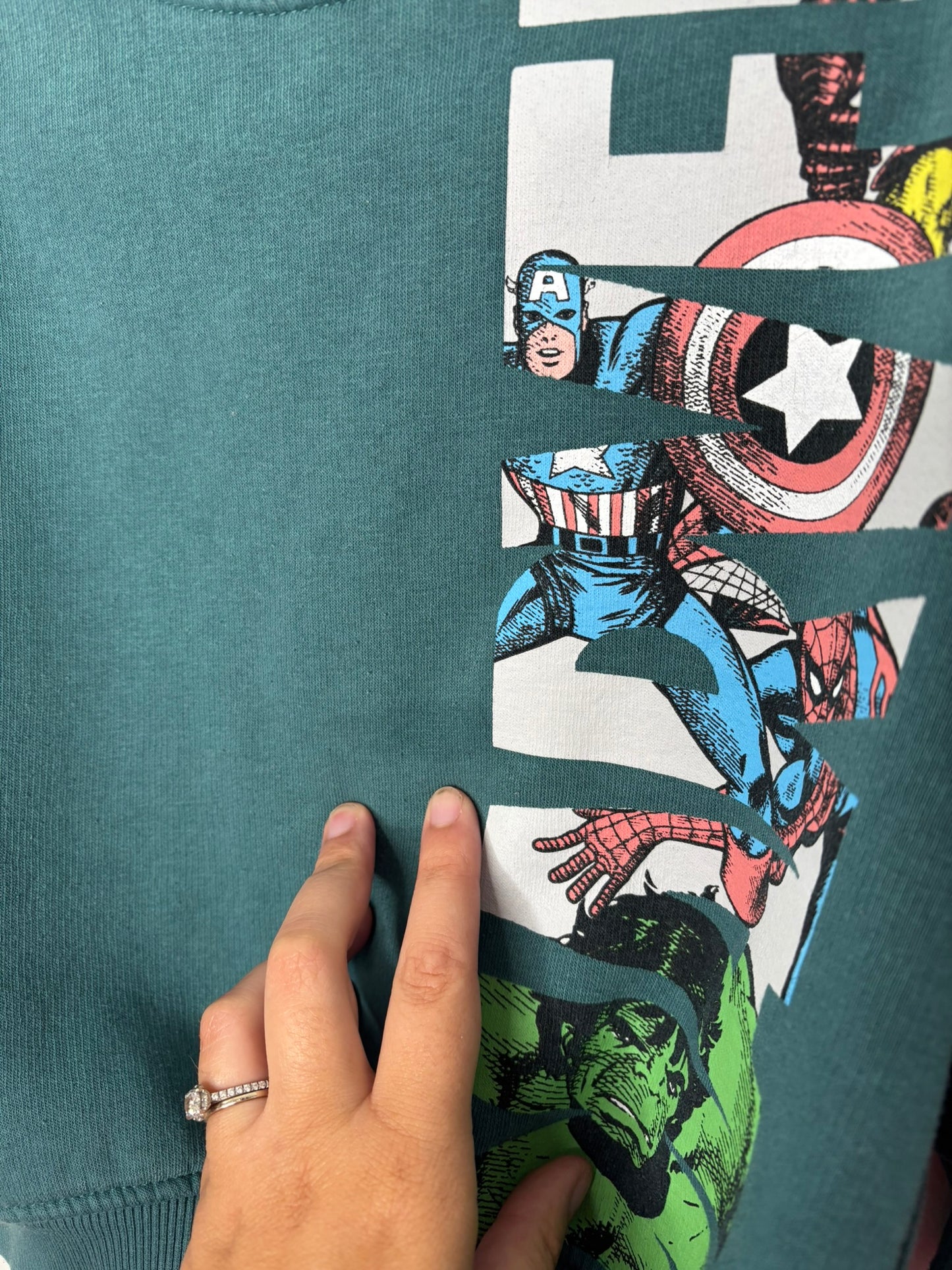 Teal Marvel Avengers sweatshirt featuring comic book characters on one side, displayed on a hanger against a white brick wall.