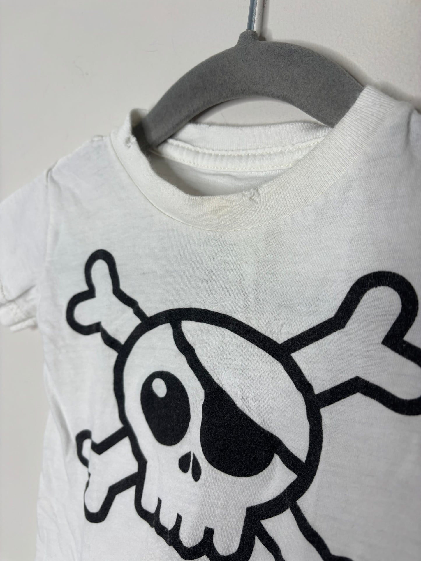 White T-shirt with a black skull and crossbones graphic, hanging on a hanger against a brick wall.