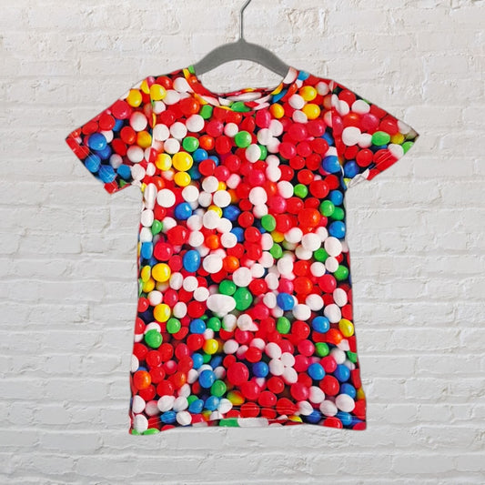 Vibrant multicolor T-shirt covered in a candy print design, displayed against a white brick wall.