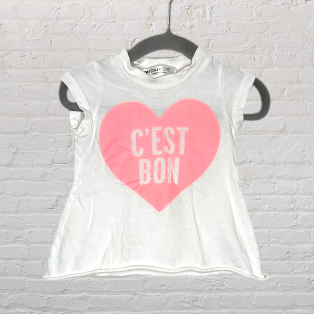 White t-shirt with a pink heart and 'C'est Bon' text, by Noah Love, hanging on a black hanger.