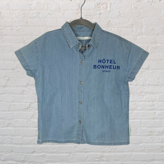 Light denim blue button-up shirt with 'Hôtel Bonheur Staff' printed on the chest, displayed on a hanger.