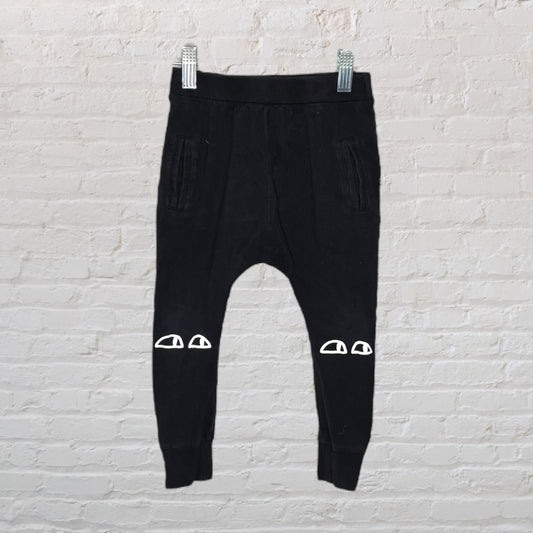Black jogger pants with white graphic details on the knees, displayed on a hanger against a white brick background.