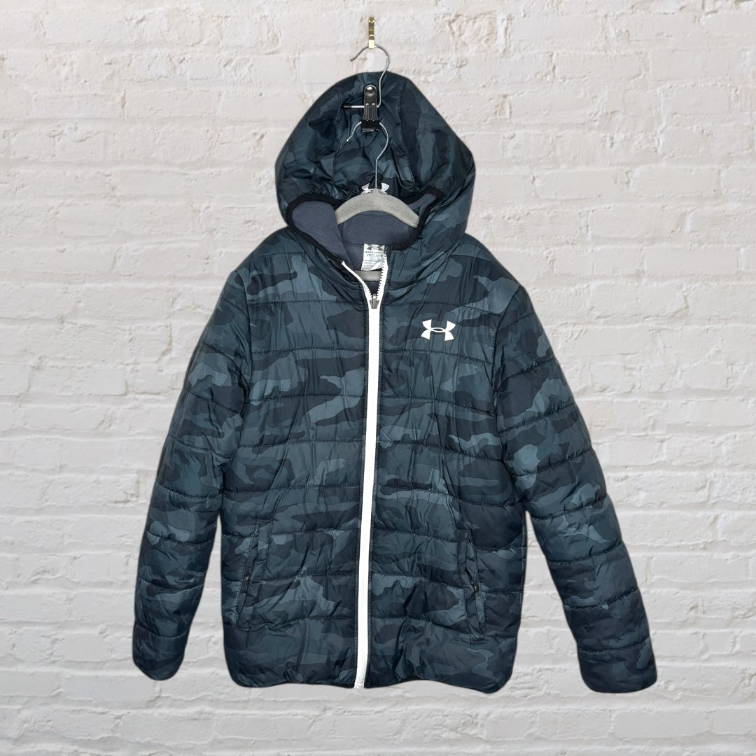 Camouflage patterned Under Armour puffer jacket with hood and white zipper, hanging on a hanger against a brick wall.
