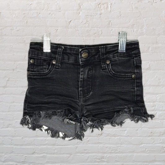 Distressed black denim shorts with frayed hems, displayed against a white brick background.