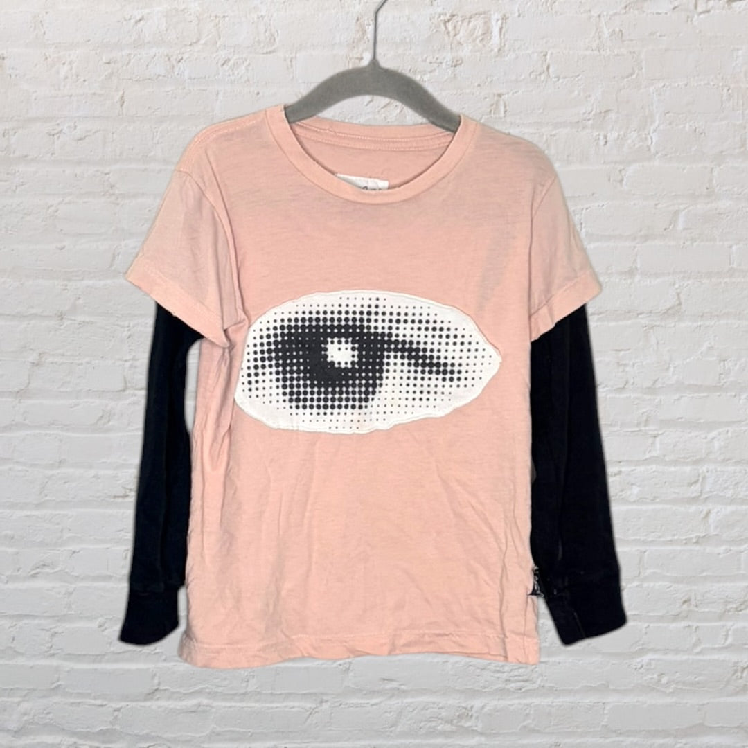Pink short sleeve T-shirt with a large black and white eye graphic, layered over black long sleeves, hanging on a hanger.