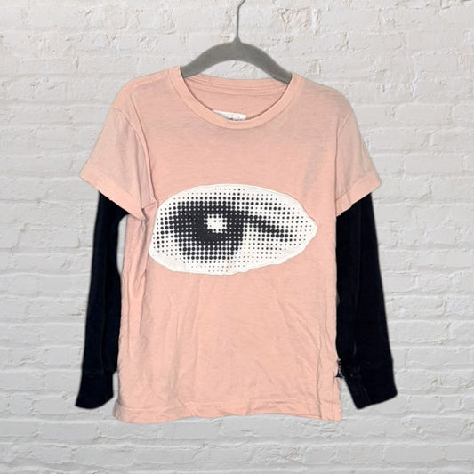 Pink short sleeve T-shirt with a large black and white eye graphic, layered over black long sleeves, hanging on a hanger.