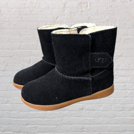 Black UGG boots with shearling lining and brown sole, side view against a brick wall background.