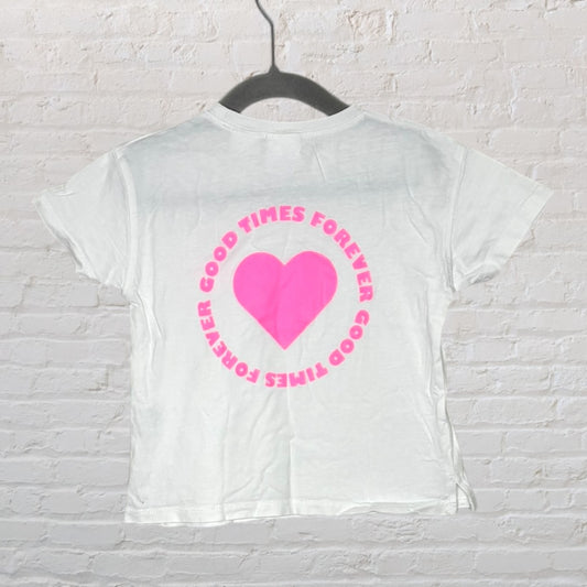 White T-shirt featuring a pink heart with 'Good Times Forever' text on the back, displayed on a hanger against a brick wall.