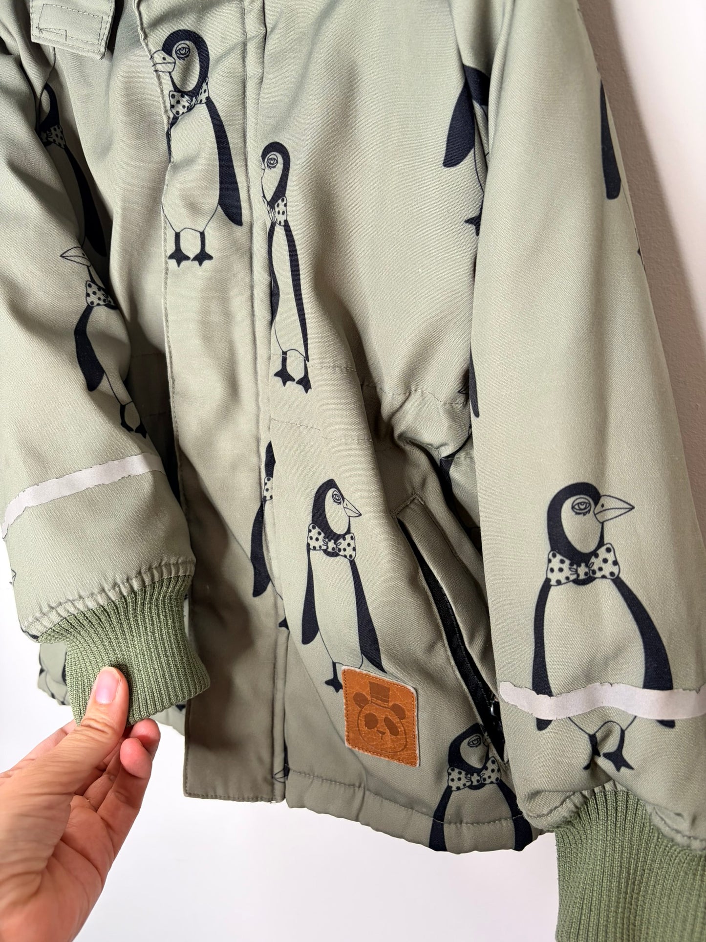 Green hooded parka with black penguin print and faux fur trim, showcased against a white brick wall.