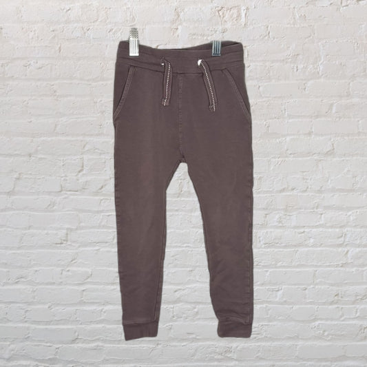 Brown jogger pants with drawstring and pockets, displayed on a hanger against a white brick wall.