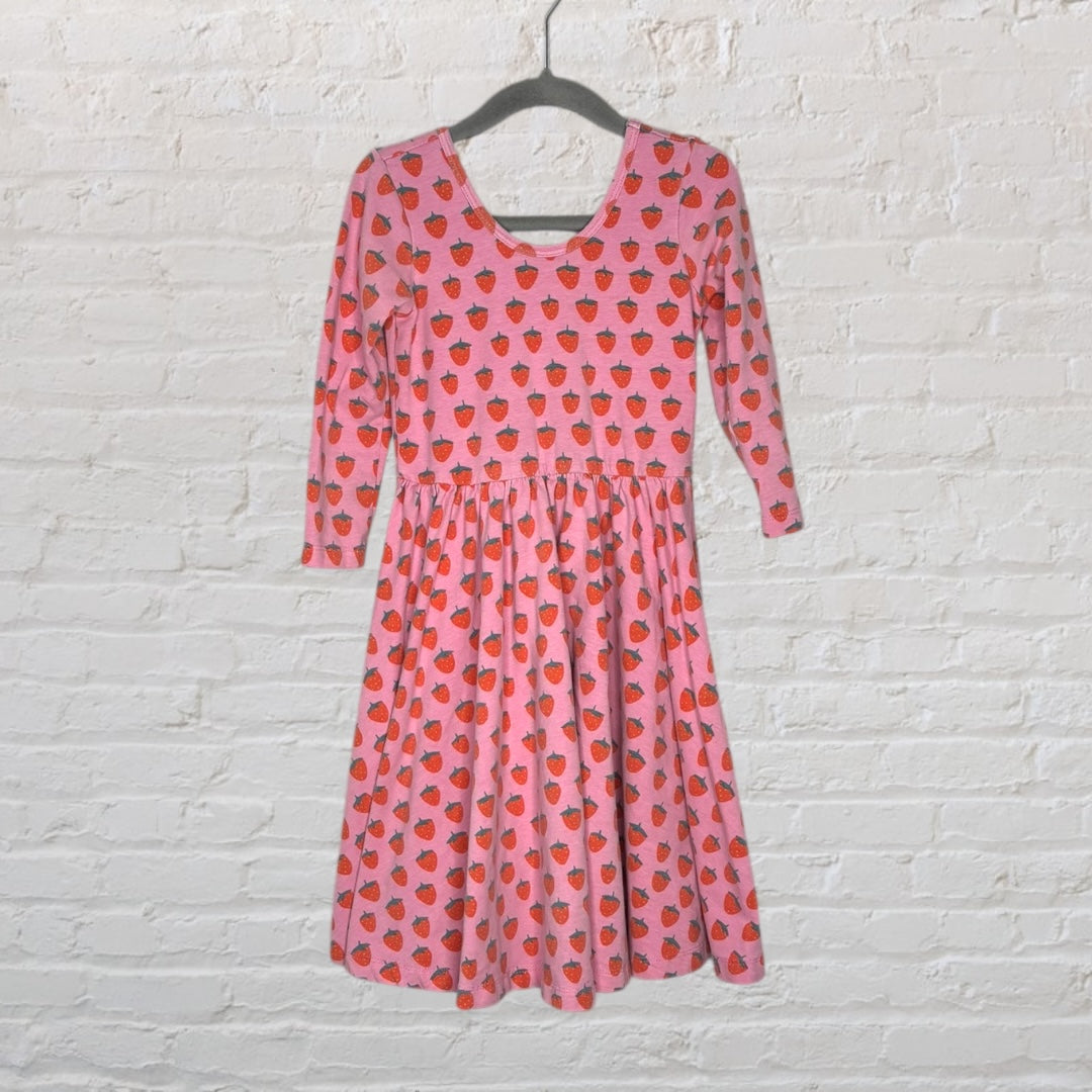 Pink skater dress with red strawberry print and three-quarter sleeves, hanging on a hanger against a white brick background.