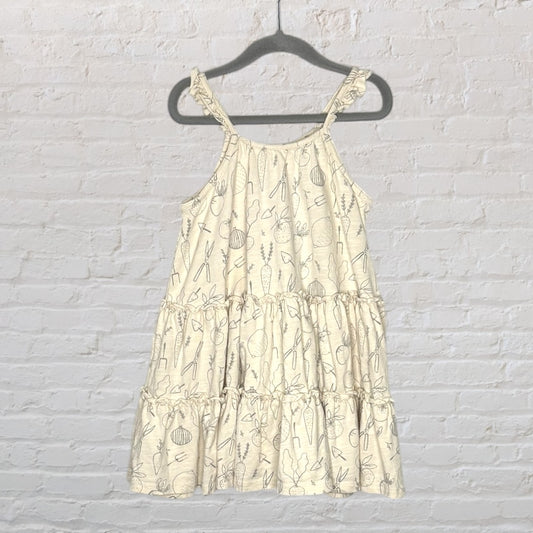 Cream tiered sleeveless dress with subtle vegetable print, hanging against a white brick wall.