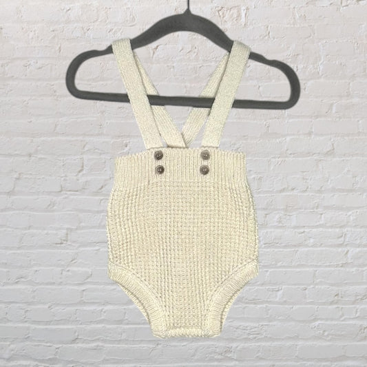 Cream knitted baby suspender romper with button details hanging on a black hanger.