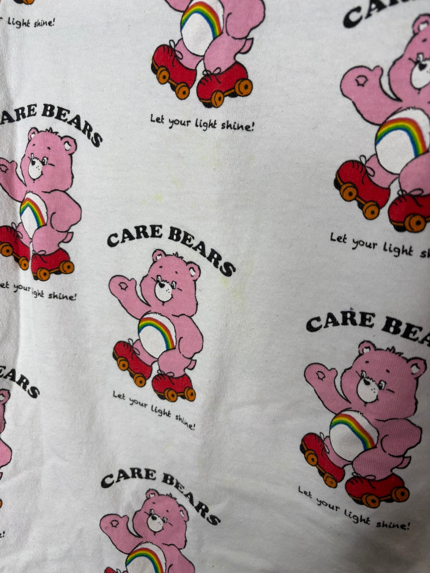 White long sleeve shirt featuring a repeating print of pink Care Bears with the text 'Let your light shine!'