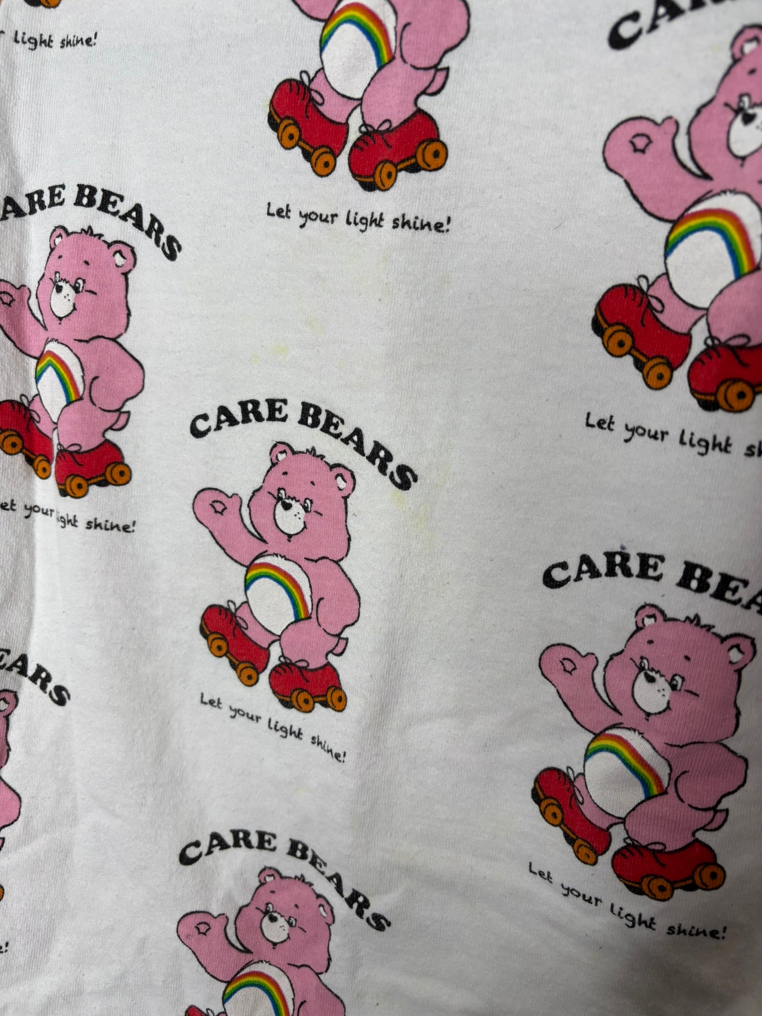 White long sleeve shirt featuring a repeating print of pink Care Bears with the text 'Let your light shine!'