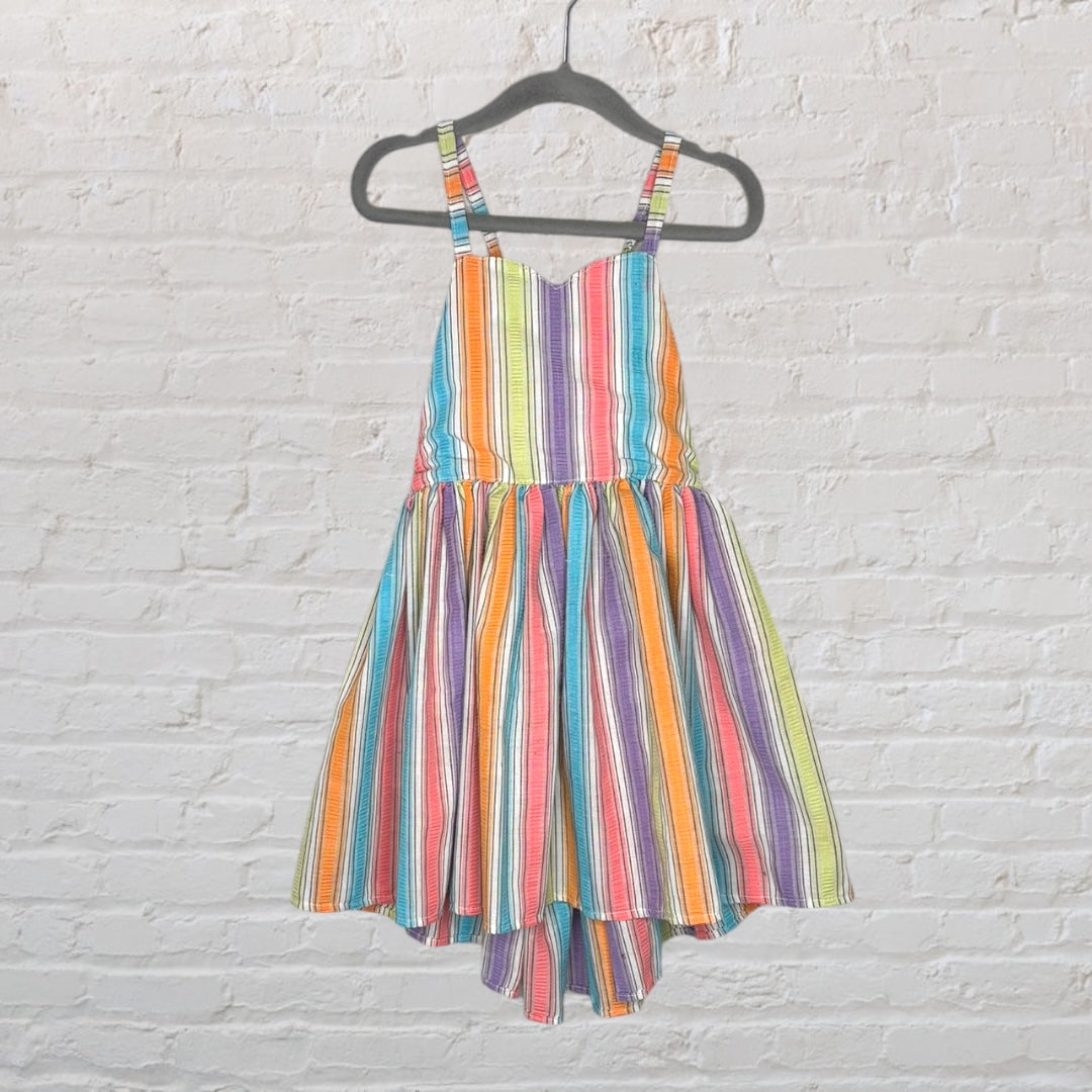 A vibrant rainbow-striped sundress with spaghetti straps, hanging on a black hanger against a white brick wall.