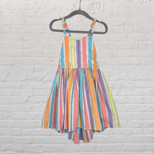A vibrant rainbow-striped sundress with spaghetti straps, hanging on a black hanger against a white brick wall.