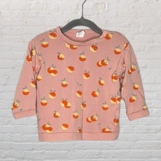 Pink toddler sweatshirt with a playful peach pattern, hanging on a hanger against a white brick wall.