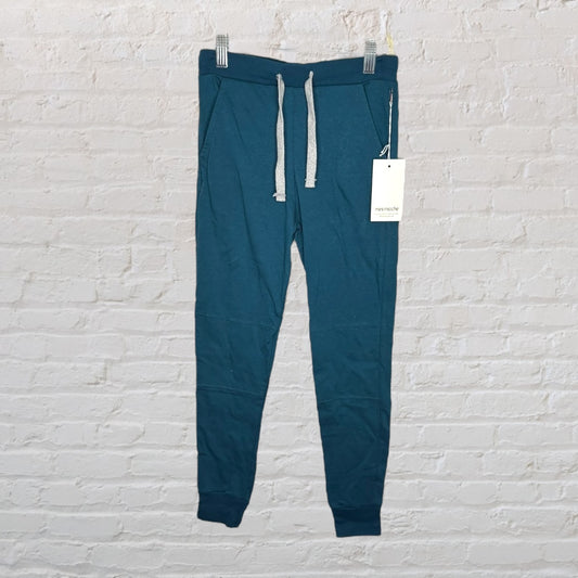 Teal jogger pants with drawstring waist and front pockets, brand Mini Mioche, hanging against a white brick wall.