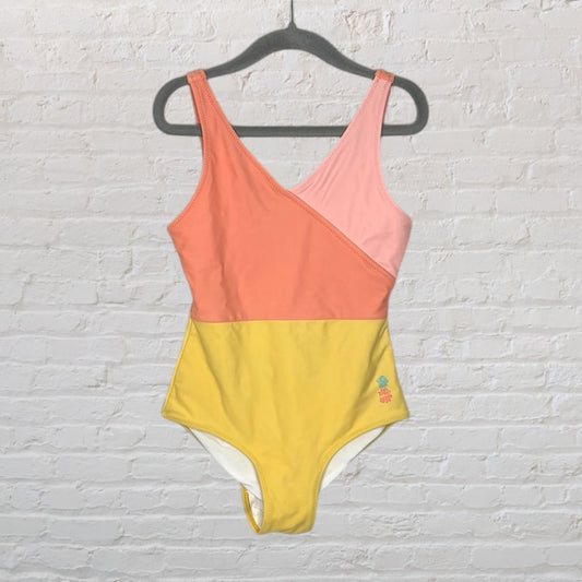 One-piece swimsuit with peach, pink, and yellow color blocks, hanging on a black hanger against a brick wall.