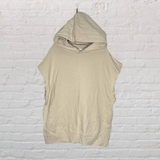 Beige sleeveless hoodie against a white brick wall.