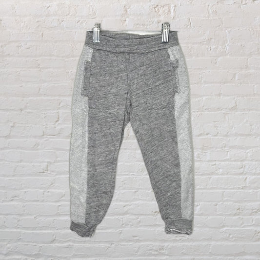 Grey jogger sweatpants with side pockets and elastic ankle cuffs, hanging on a hanger against a brick background.