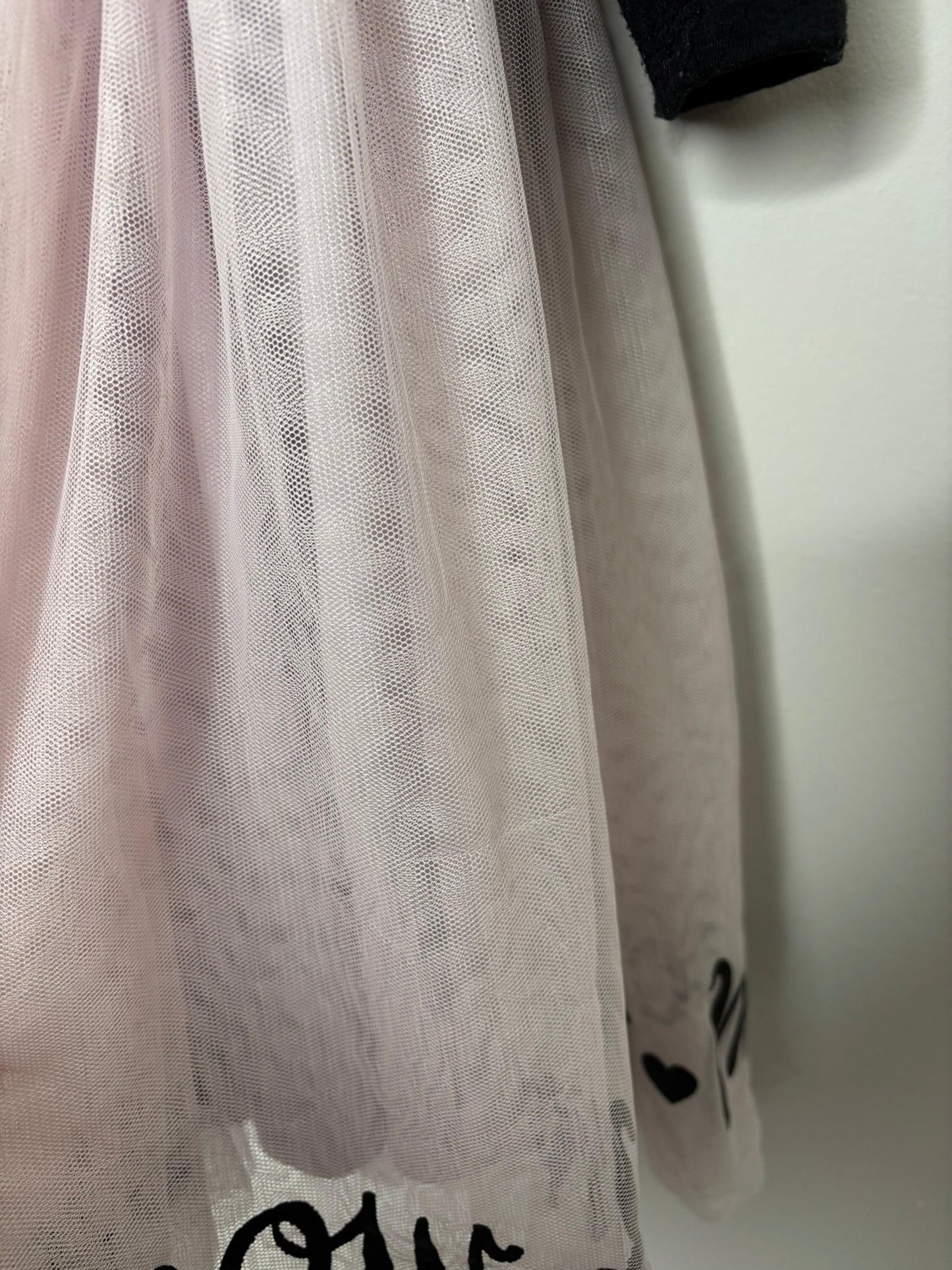 Black long sleeve dress with cat face design and pink tulle skirt, hanging on a hanger against a white brick wall.