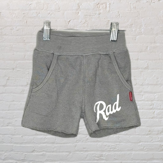 Grey shorts with 'Rad' graphic text on the leg, displayed against a white brick wall.