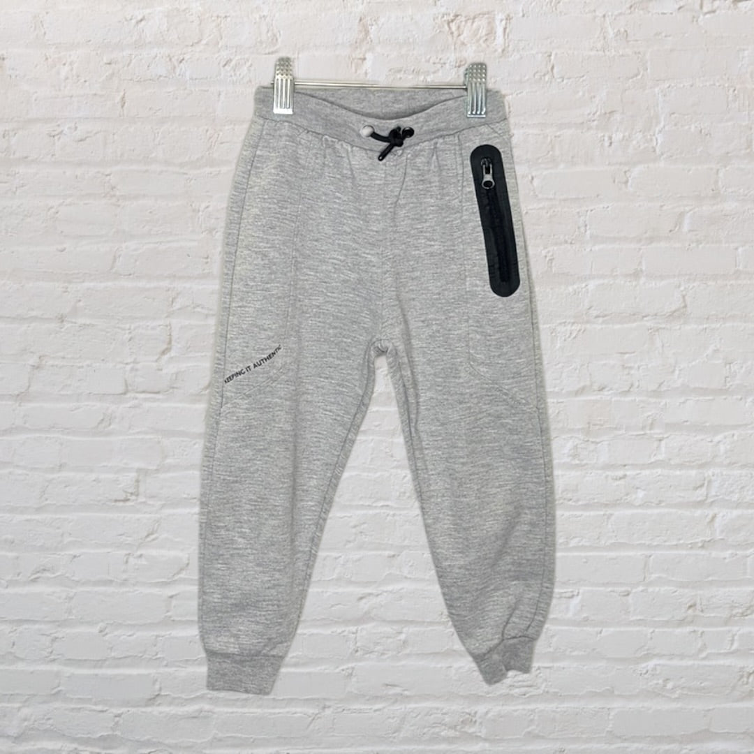 Grey jogger sweatpants with a black zipper pocket and drawstring, hanging on a white brick wall.