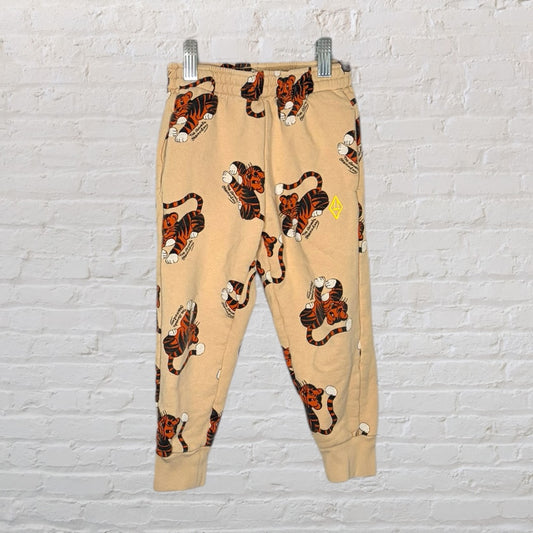 Beige jogger pants with an all-over tiger print design, displayed on hangers against a white brick wall.