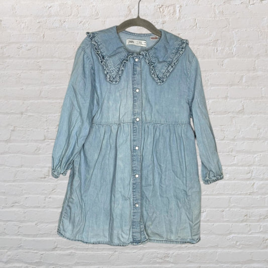 Light blue denim button-down dress with a ruffled collar and long sleeves, hanging on a hanger against a white brick wall.