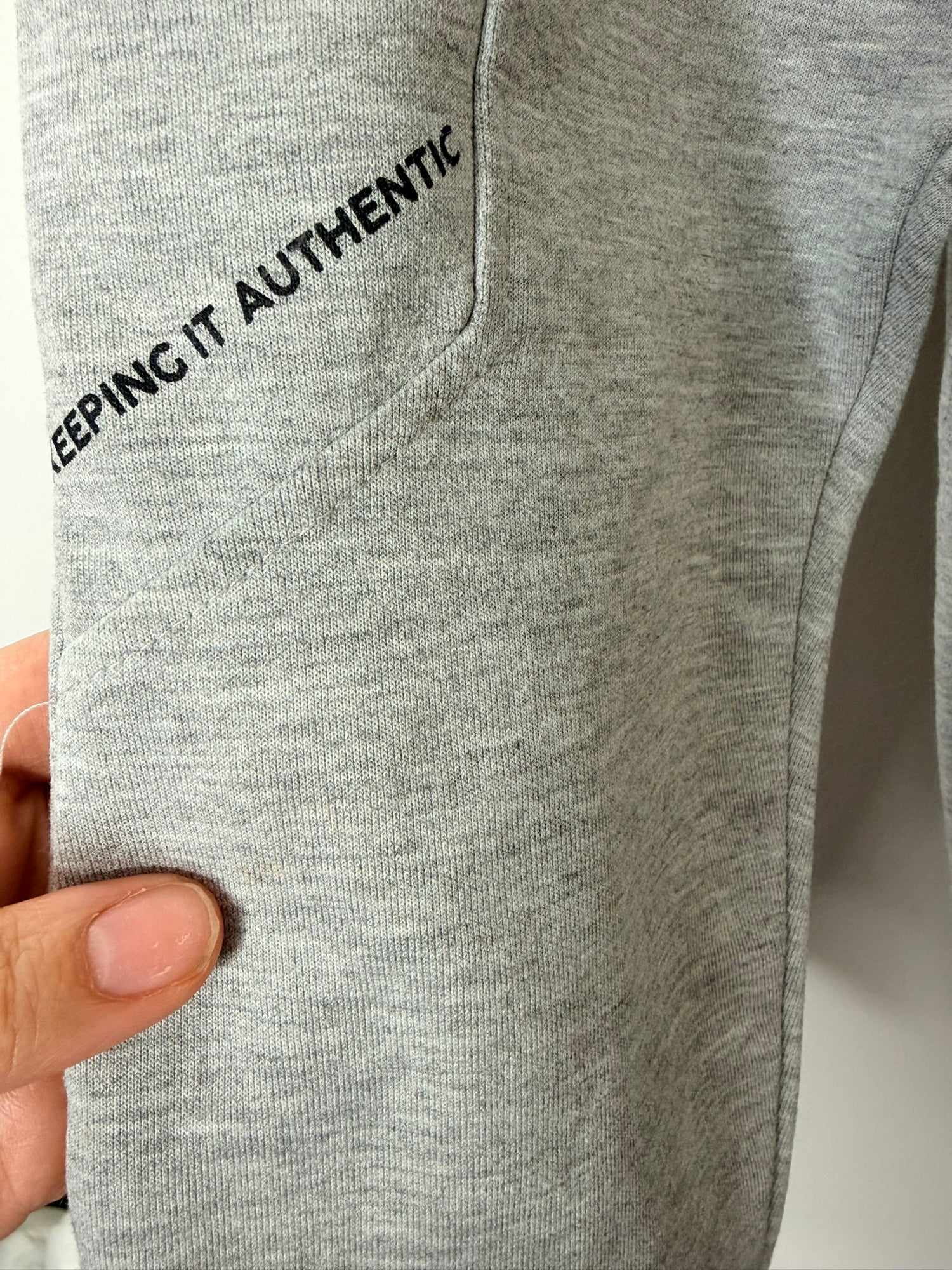 Grey jogger sweatpants with a black zipper pocket and drawstring, hanging on a white brick wall.